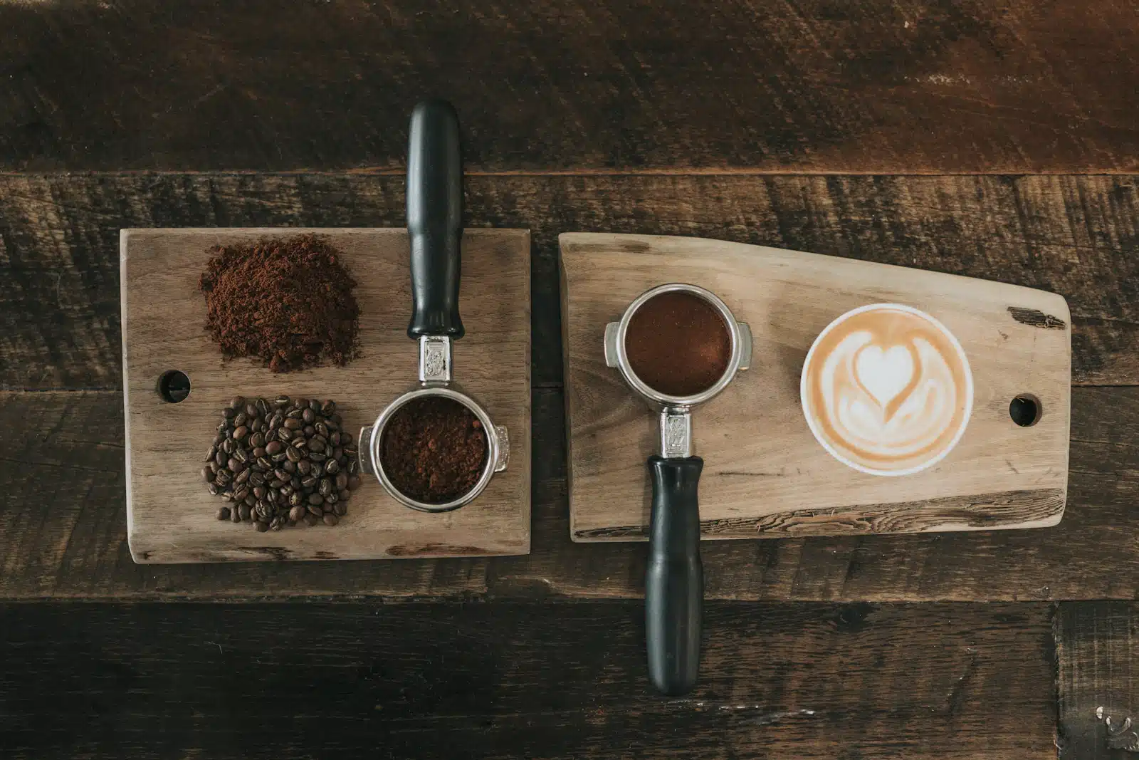 Granos de café tostados junto a café molido y portafiltros sobre tablas de madera, al lado de un latte