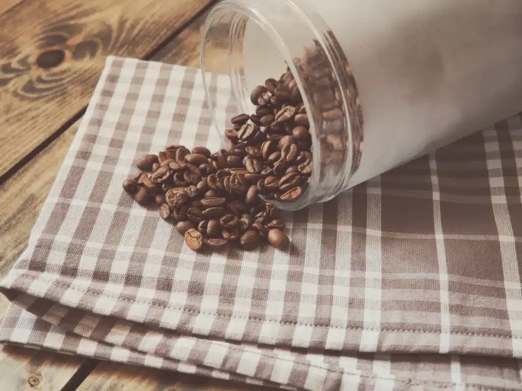 Frasco de vidrio volcado sobre un mantel de cuadros, con granos de café tostado regados sobre la mesa