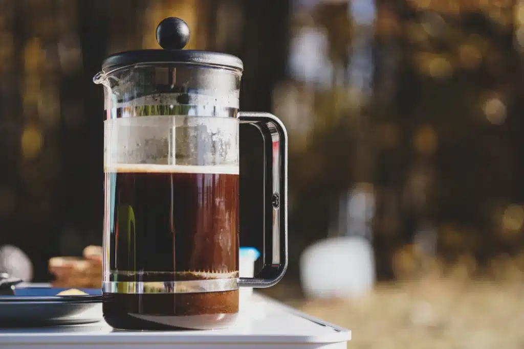 Prensa francesa de vidrio con café recién preparado sobre una mesa, con luz cálida de fondo