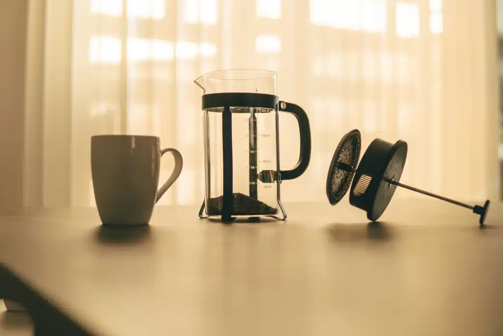 Prensa francesa de vidrio con café molido grueso en el fondo y el émbolo aparte, junto a una taza blanca