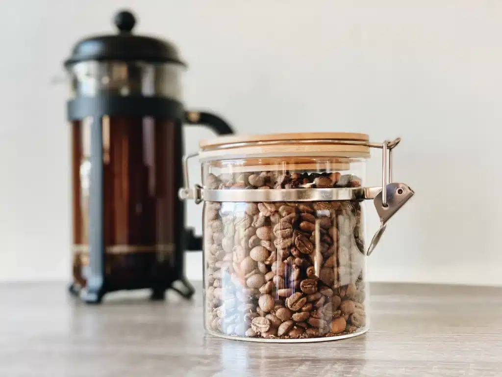 Prensa francesa con café recién preparado junto a un frasco de vidrio lleno de granos de café tostados