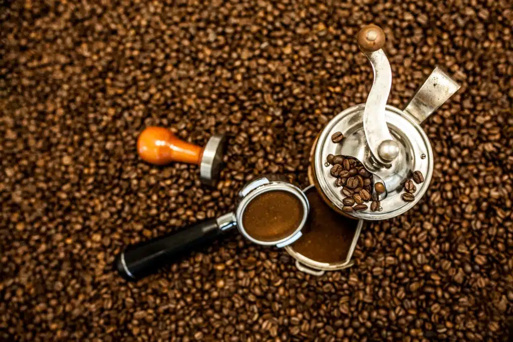 Molino manual de café sobre granos tostados, junto a un portafiltro con café molido fino para espresso