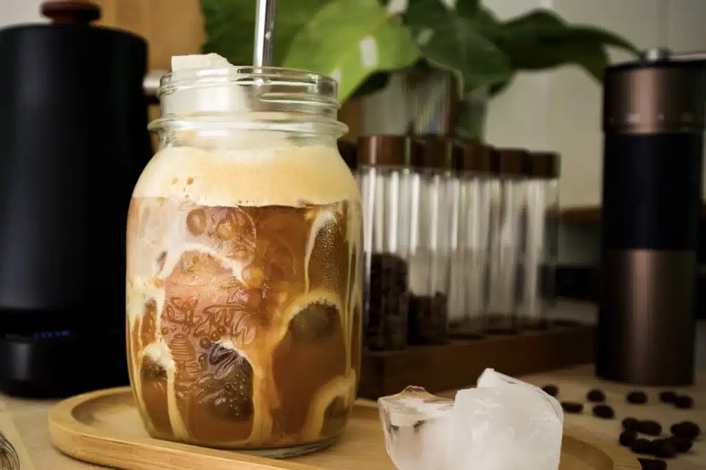 Frasco de vidrio con cold brew, hielo y leche, junto a un molino de café y granos tostados sobre una bandeja de madera