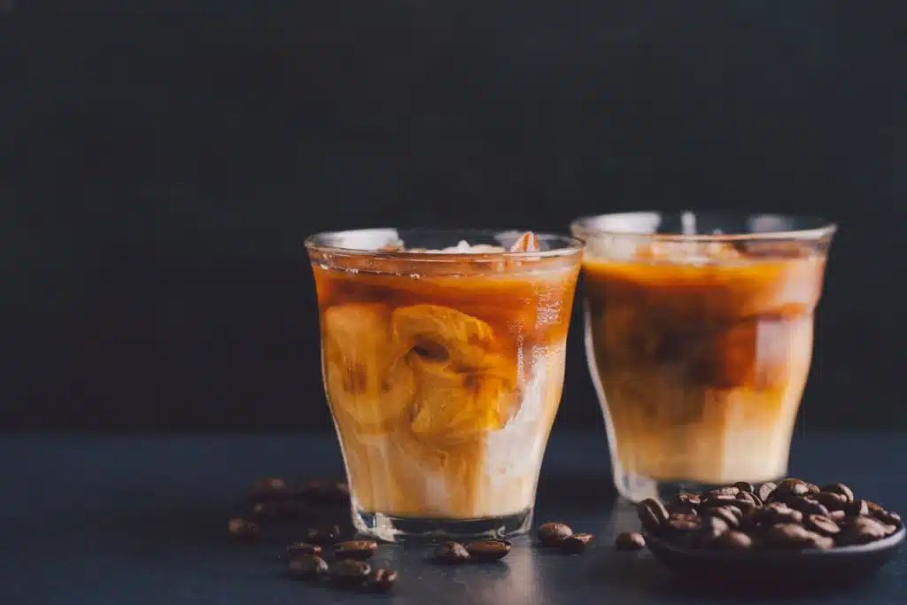 Dos vasos de café frío con leche y hielo sobre una mesa oscura, rodeados de granos de café tostado