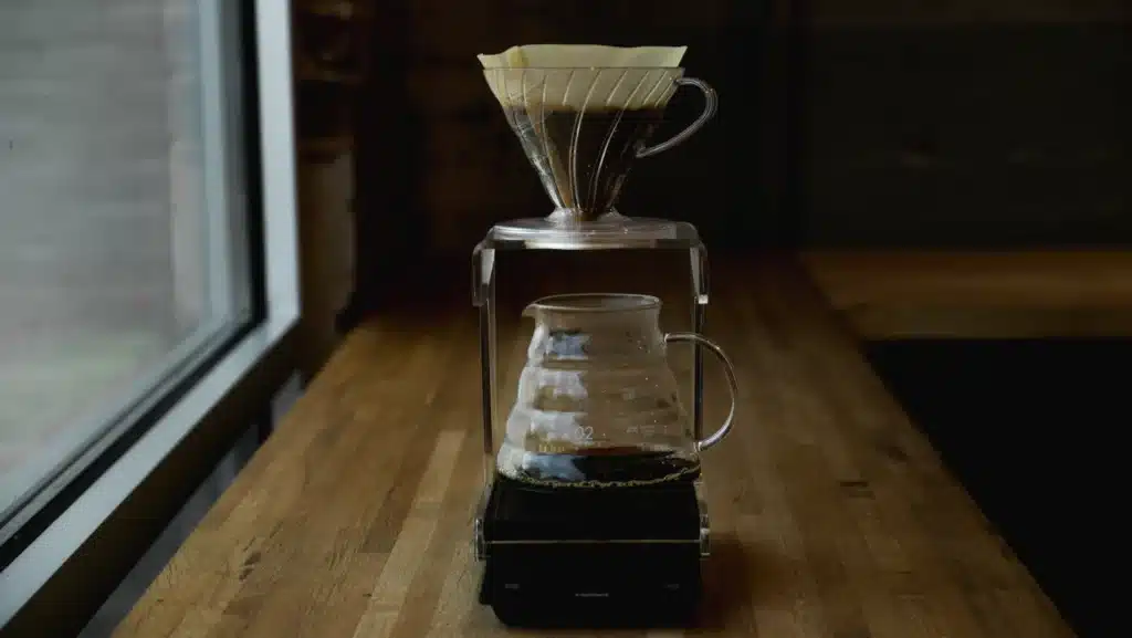 Gotero V60 con filtro de papel sobre una jarra de vidrio y una balanza, junto a una ventana