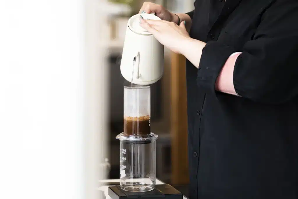 Persona vertiendo agua caliente con un hervidor en una AeroPress puesta sobre una jarra de vidrio y una balanza