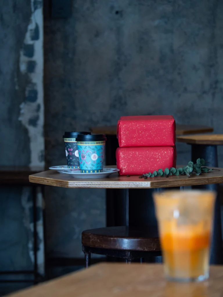Dos vasos de café de papel estampado junto a dos cajas de regalo rojas sobre una mesa de madera